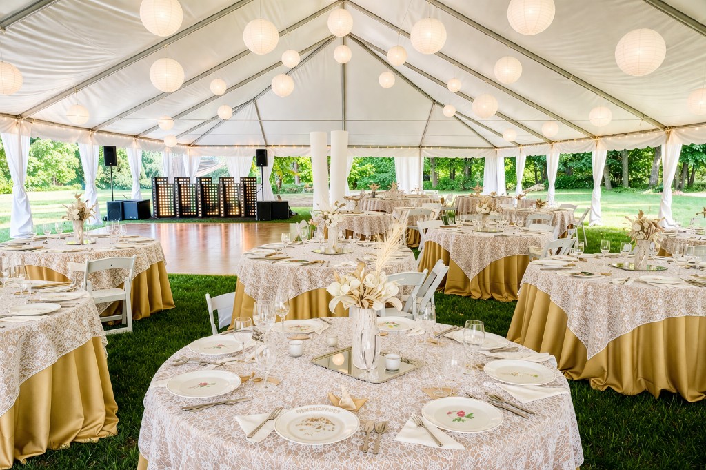 Outdoor wedding reception under a white frame tent, champagne gold linens, white lanterns, string lights, dance floor, and lawn beyond.